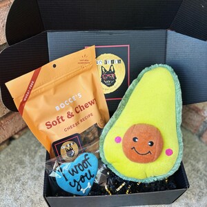 May include: A black gift box filled with dog treats and toys. The box contains a bag of "Bocce's Soft & Chewy Cheese Recipe" dog treats, a blue heart-shaped dog biscuit that says "I woof you", and a plush avocado-shaped dog toy.