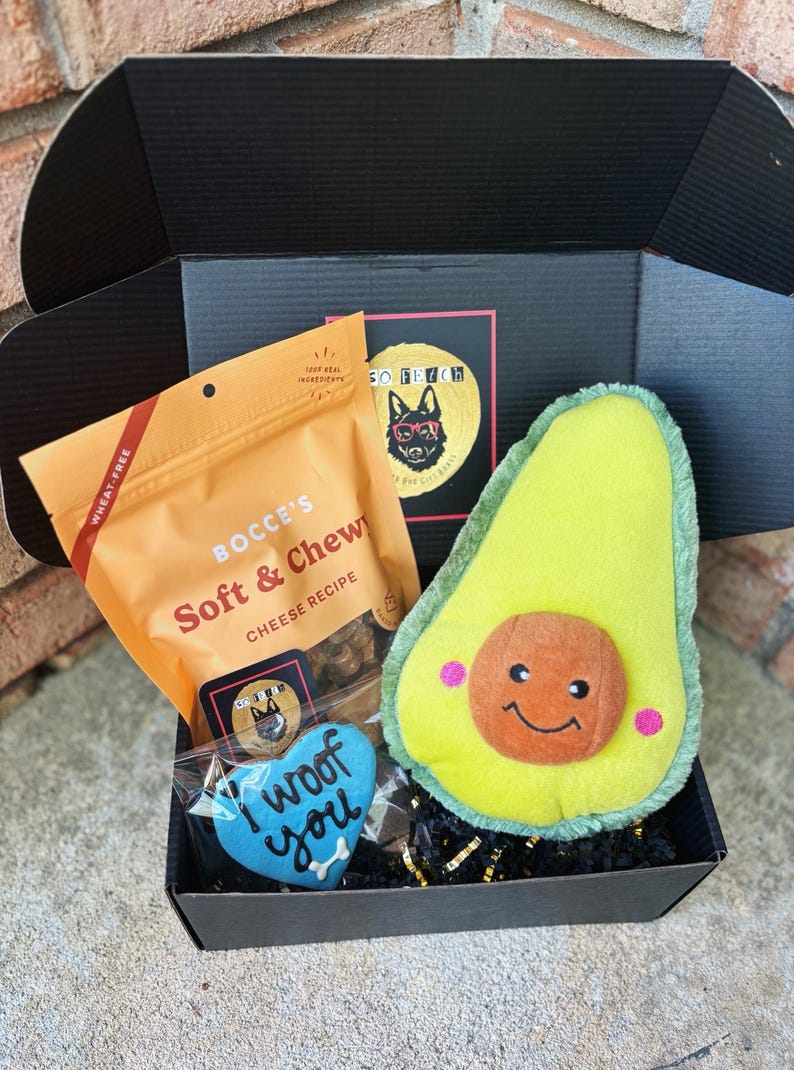 May include: A black gift box filled with dog treats and toys. The box contains a bag of "Bocce's Soft & Chewy Cheese Recipe" dog treats, a blue heart-shaped dog biscuit that says "I woof you", and a plush avocado-shaped dog toy.