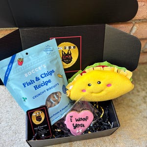 May include: A black gift box filled with dog treats and toys. The box contains a bag of "Fish & Chips Recipe" crunchy biscuits, a plush yellow taco toy, and a pink heart-shaped cookie that says "I woof you".