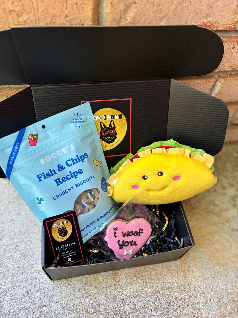 May include: A black gift box filled with dog treats and toys. The box contains a bag of "Fish & Chips Recipe" crunchy biscuits, a plush yellow taco toy, and a pink heart-shaped cookie that says "I woof you".
