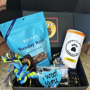 May include: A blue and black gift box filled with dog treats, a blue rope toy, a blue heart-shaped cookie with the text "I woof you", and a white can-shaped plush toy with the text "Zippy Claw Spiked Seltzer Mango".