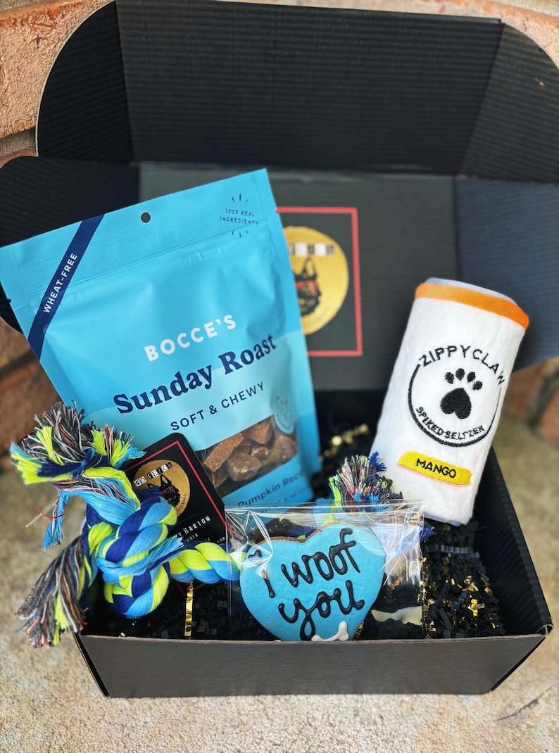 May include: A blue and black gift box filled with dog treats, a blue rope toy, a blue heart-shaped cookie with the text "I woof you", and a white can-shaped plush toy with the text "Zippy Claw Spiked Seltzer Mango".