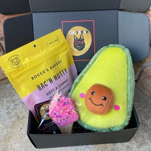 May include: A black gift box filled with dog treats and toys. The box contains a bag of "Bac'n Nutty" dog treats, a pink and white frosted dog treat, and a plush avocado-shaped dog toy with a smiling face.