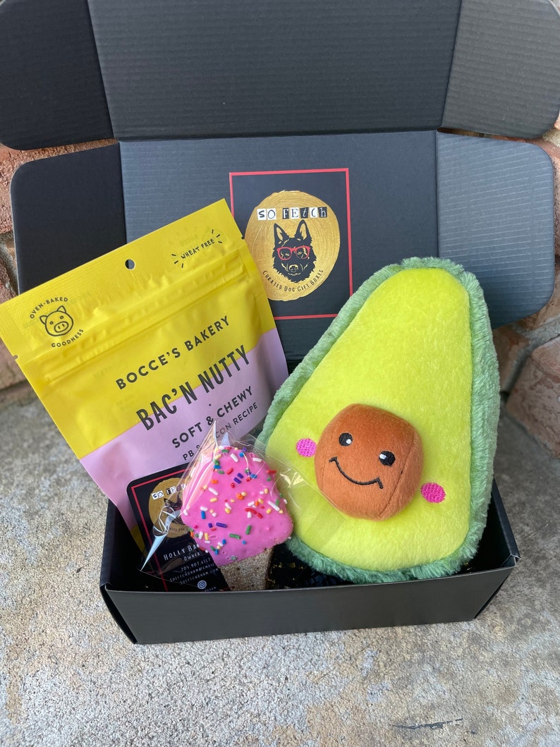 May include: A black gift box filled with dog treats and toys. The box contains a bag of "Bac'n Nutty" dog treats, a pink and white frosted dog treat, and a plush avocado-shaped dog toy with a smiling face.