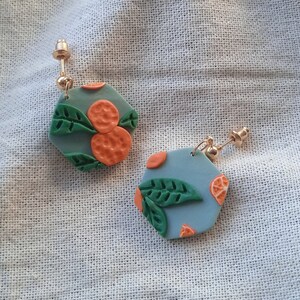 Könnte beinhalten: Ein Paar sechseckige Ohrringe mit blauem Hintergrund und orangefarbenen und grünen Designs. Die Ohrringe sind aus Polymer Clay gefertigt und haben goldene Ohrstecker.