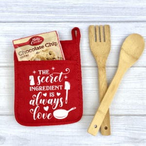 May include: Red kitchen towel with a pocket that says "The secret ingredient is always love". A packet of chocolate chip cookie mix is in the pocket. Two wooden kitchen utensils are on a white wood surface.