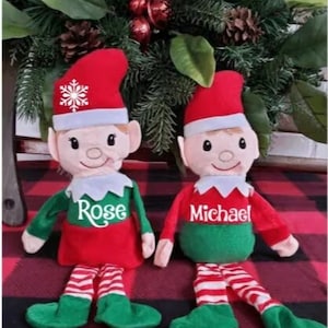 Può includere: Due bambole di elfo di peluche con abiti rossi e verdi. L'elfo a sinistra indossa un vestito rosso con strisce bianche e l'elfo a destra indossa una camicia verde con strisce bianche. Entrambi gli elfi hanno fiocchi di neve bianchi sui loro cappelli e sono personalizzati con i nomi "Rose" e "Michael".