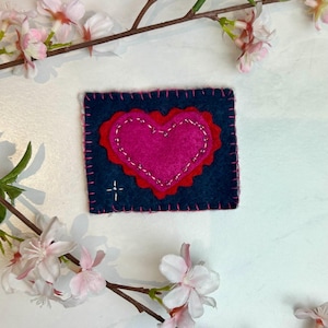 May include: A handmade felt heart-shaped patch. The patch features a dark blue background with a red scalloped heart and a bright pink heart in the center. The edges are stitched with pink thread, and a small cross is stitched in the corner.
