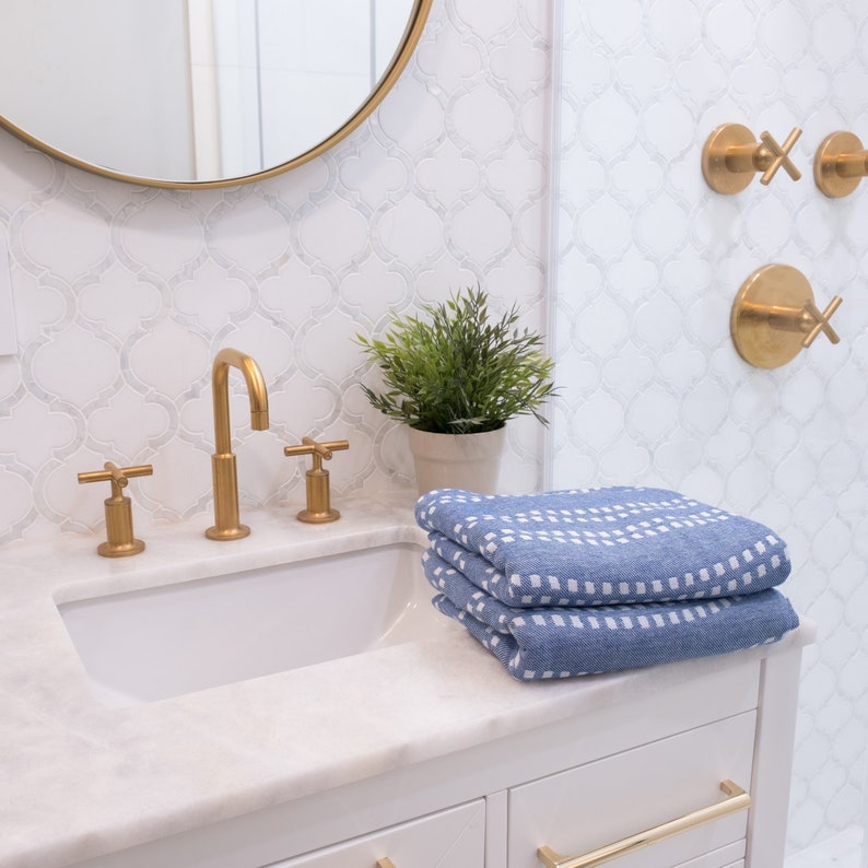 May include: A white bathroom vanity with a white porcelain sink and gold faucets. Three blue towels with white geometric patterns are stacked on the counter. The bathroom has white subway tile with a decorative pattern.