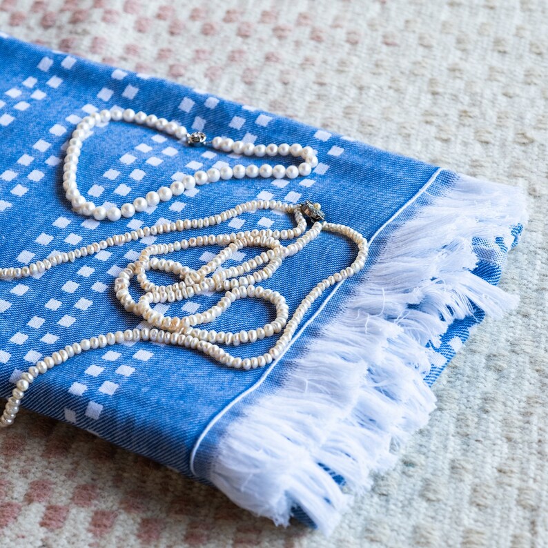 May include: Two pearl necklaces on a blue and white patterned fabric with white fringe. The fabric has a checkered pattern.