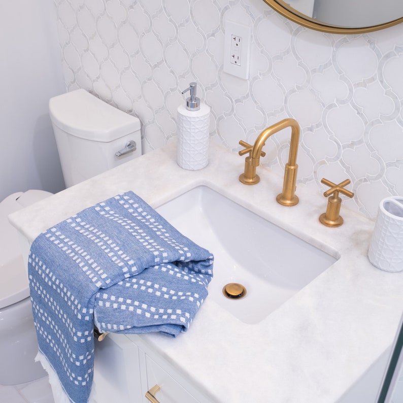 May include: A white bathroom vanity with a white porcelain sink and a gold faucet. A blue and white striped towel is draped over the sink. A white ceramic soap dispenser is on the counter.