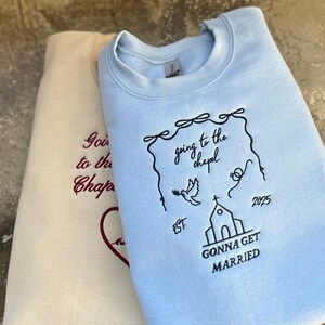 May include: Two light blue and cream colored sweatshirts with embroidered text. The light blue sweatshirt has the text "going to the chapel" and "gonna get married" with a church and dove design. The cream colored sweatshirt has the text "going to the chapel" in red.