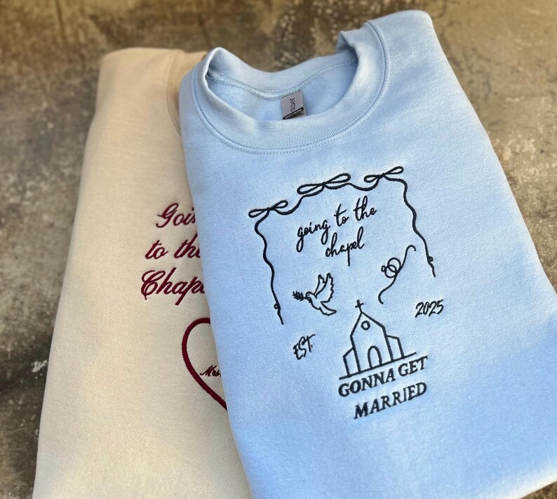 May include: Two light blue and cream colored sweatshirts with embroidered text. The light blue sweatshirt has the text "going to the chapel" and "gonna get married" with a church and dove design. The cream colored sweatshirt has the text "going to the chapel" in red.