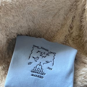 May include: Light blue sweatshirt with a black embroidered design. The design features a church, a bird, and the text "going to the chapel est 2025 gonna get married".