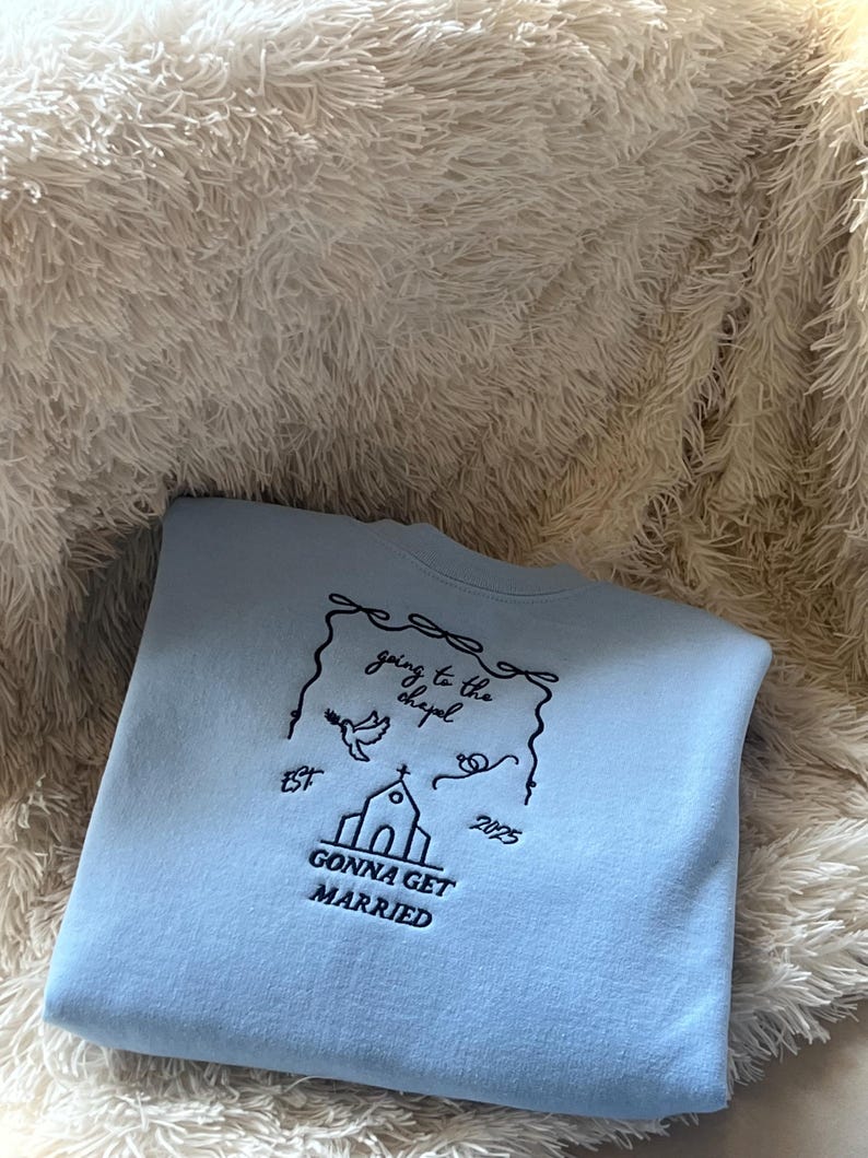 May include: Light blue sweatshirt with a black embroidered design. The design features a church, a bird, and the text "going to the chapel est 2025 gonna get married".