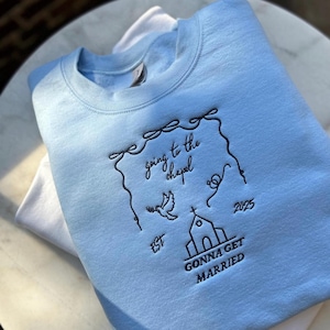 May include: Light blue sweatshirt with black embroidered text that reads "going to the chapel 2025 est. gonna get married". The embroidery includes a church, a dove, and a heart.