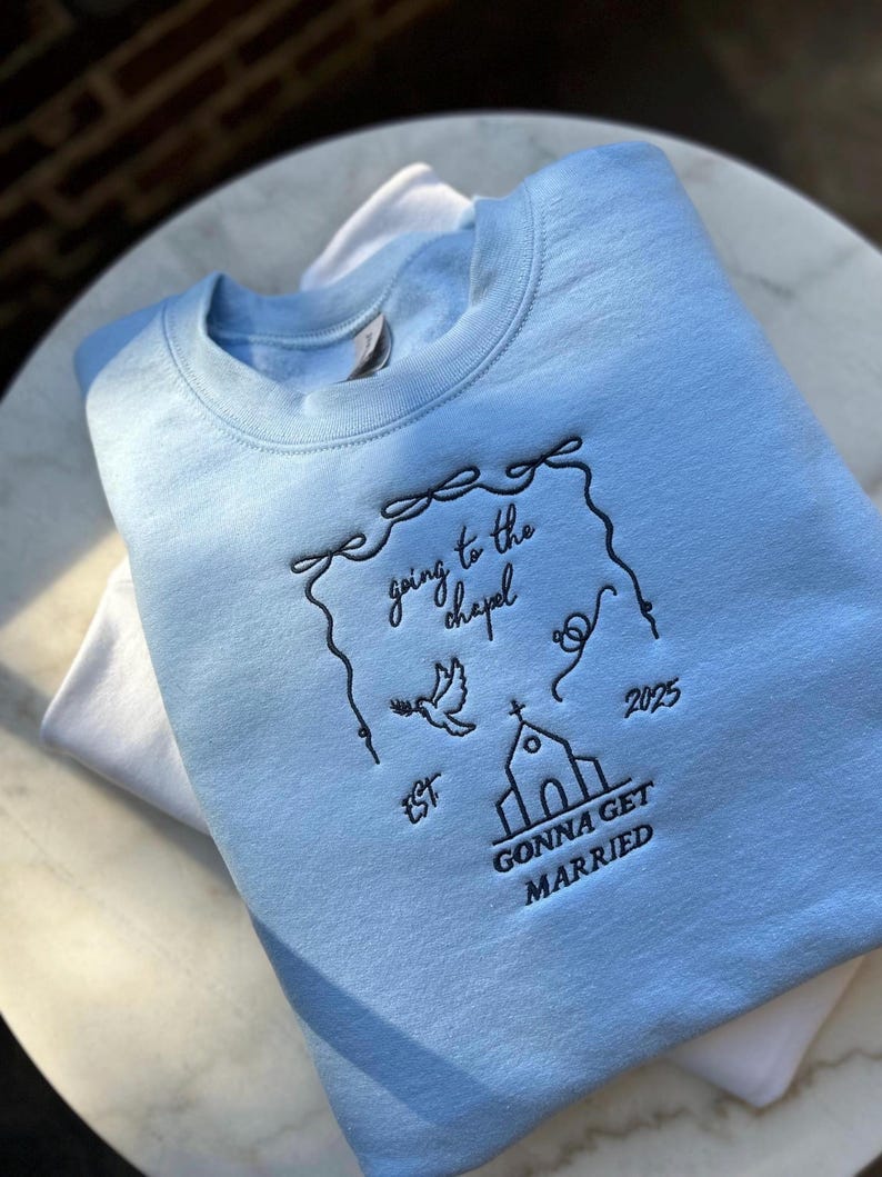 May include: Light blue sweatshirt with black embroidered text that reads "going to the chapel 2025 est. gonna get married". The embroidery includes a church, a dove, and a heart.