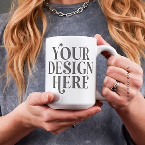 May include: A white ceramic mug with a grey inscription that reads "YOUR DESIGN HERE". The mug is being held by a person wearing a grey shirt.