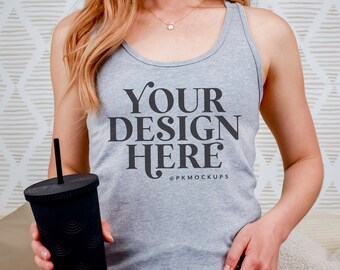 May include: A woman wearing a gray tank top with the text "YOUR DESIGN HERE" and the copyright symbol "©PKMOCKUPS". She is holding a black reusable cup with a straw. She is wearing denim shorts.