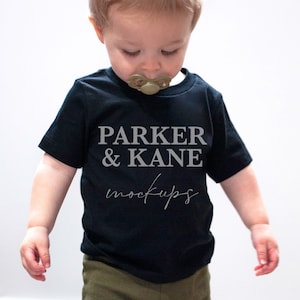 Pu&ograve; includere: Un bambino piccolo che indossa una maglietta nera con il testo "PARKER & KANE mockups" in lettere grigie. Il bambino indossa pantaloni verde oliva e guarda in basso.