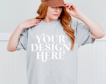 May include: A woman wearing a gray t-shirt with the text "YOUR DESIGN HERE" printed on the front. She is wearing a brown baseball cap and has long red hair.