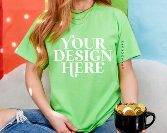 May include: A woman wearing a bright green t-shirt with the text "YOUR DESIGN HERE" in white. She is sitting on a white surface with a rainbow backdrop and holding a black mug filled with gold coins.