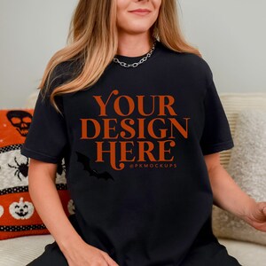 May include: A black t-shirt with the text "YOUR DESIGN HERE" in orange lettering and a small bat graphic. The t-shirt is being worn by a person sitting on a couch.