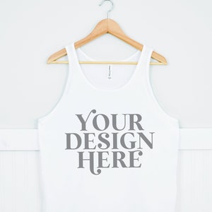 May include: White tank top with a wooden hanger. The tank top has the text "YOUR DESIGN HERE" printed on the front in gray.