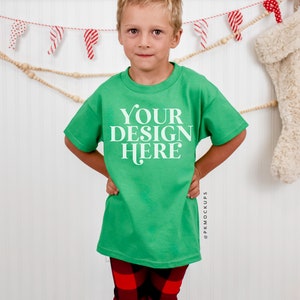 May include: A young person wearing a green t-shirt with the text "YOUR DESIGN HERE" in white. The person is standing in front of a white wall with a garland of red and white candy canes hanging above.