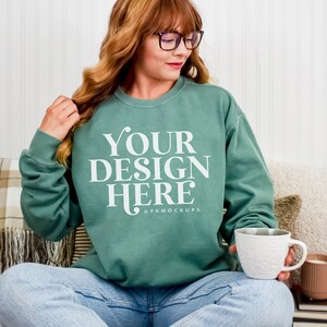 May include: A teal sweatshirt with the text "YOUR DESIGN HERE" in white. The person is wearing glasses and holding a white mug. A candle and book are on a table. The sweatshirt is a mock-up for a design.