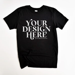 May include: A black t-shirt with the text "YOUR DESIGN HERE" in white lettering and the text "@PKMOCKUPS" in smaller white lettering below.
