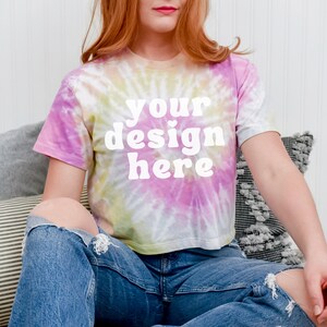 May include: A tie-dye crop top with a white "your design here" text in a retro font. The top is worn by a person sitting on a couch with pillows.