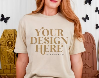 May include: A beige short-sleeved t-shirt featuring the dark brown text "YOUR DESIGN HERE" and "@PKMOCKUPS" printed on the front. The shirt is worn by a person with long reddish-brown hair, positioned against a backdrop of decorative tombstones.