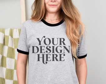May include: A gray ringer t-shirt with black trim around the collar and sleeves. The text "YOUR DESIGN HERE" is printed in black on the front. The model is wearing blue jeans.