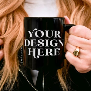 May include: A black ceramic mug with the text "YOUR DESIGN HERE" in white. The mug is held by a person. The mug has a handle and is a standard size for coffee or tea.