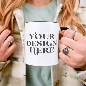 May include: White ceramic mug with a black rim and handle. The mug has the text "YOUR DESIGN HERE" printed on the front.
