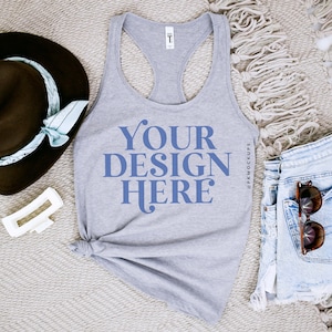 May include: A gray tank top with the text "YOUR DESIGN HERE" in blue. The tank top is laying on a white fuzzy surface with a brown hat, a white hair clip, and a pair of blue denim shorts with sunglasses in the pocket.