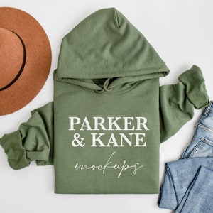 May include: A green hooded sweatshirt with the text "PARKER & KANE mockups" printed on the front. The sweatshirt is laid flat on a white surface with a brown hat and blue jeans.