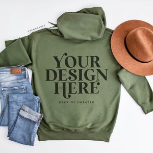 May include: An olive green hoodie with the text "YOUR DESIGN HERE" in black, paired with light blue jeans and a brown hat. The back of the sweater is visible, showcasing the design space. The hat is a warm brown color.