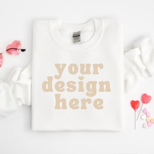 Op de afbeelding: Witte trui met de tekst "your design here" in een lichtbruine kleur. De trui ligt plat op een wit oppervlak met roze hartvormige zonnebrillen en rode en roze hartvormige lolly's.