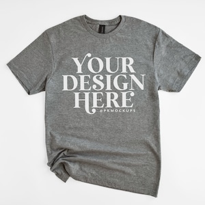 May include: A heather grey t-shirt with short sleeves and a crew neck. The shirt has the text "YOUR DESIGN HERE" printed on the front in white.