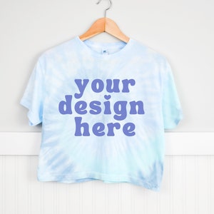 Pode incluir: Uma camiseta cropped azul claro tie-dye com mangas curtas pendurada em um cabide de madeira. A camiseta tem o texto "your design here" impresso em roxo na frente.