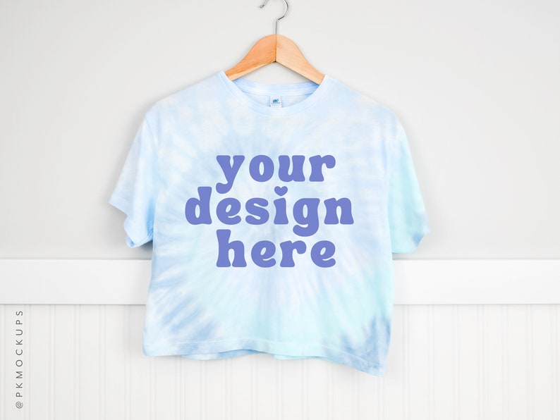 Pode incluir: Uma camiseta cropped azul claro tie-dye com mangas curtas pendurada em um cabide de madeira. A camiseta tem o texto "your design here" impresso em roxo na frente.