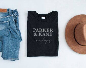 May include: A black t-shirt with the text "Parker & Kane mockups" printed on the front. The t-shirt is folded and laid flat on a white surface. There are also a pair of blue jeans and a brown fedora hat in the image.