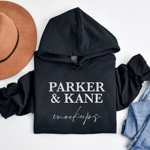 May include: Black hooded sweatshirt with the text "PARKER & KANE mockups" printed on the front. The sweatshirt is laid flat on a white surface with a brown hat and blue jeans.