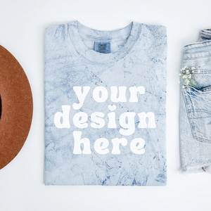 Puede incluir: Una camiseta azul claro con un patrón de tie-dye blanco. La camiseta está doblada y colocada sobre una superficie blanca. El texto "your design here" está impreso en blanco en la parte delantera de la camiseta.