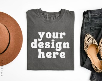 May include: A gray t-shirt with the text "your design here" in white. The t-shirt is folded and laid flat on a white surface. There is a brown hat, a pair of jeans, and a pair of leopard print shoes in the background.