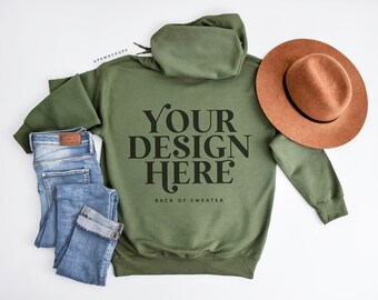 May include: A green hooded sweatshirt with the text "YOUR DESIGN HERE" and "BACK OF SWEATER" printed on the back. The sweatshirt is laid flat on a white surface with a pair of blue jeans and a brown hat.