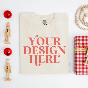 May include: A white t-shirt with the text "YOUR DESIGN HERE" in red. The t-shirt is laying flat on a white surface with various Christmas decorations around it, including red ornaments, wooden nutcrackers, and a red and white checkered gift box.