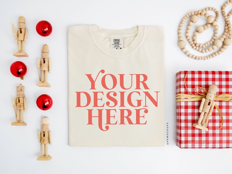 May include: A white t-shirt with the text "YOUR DESIGN HERE" in red. The t-shirt is laying flat on a white surface with various Christmas decorations around it, including red ornaments, wooden nutcrackers, and a red and white checkered gift box.